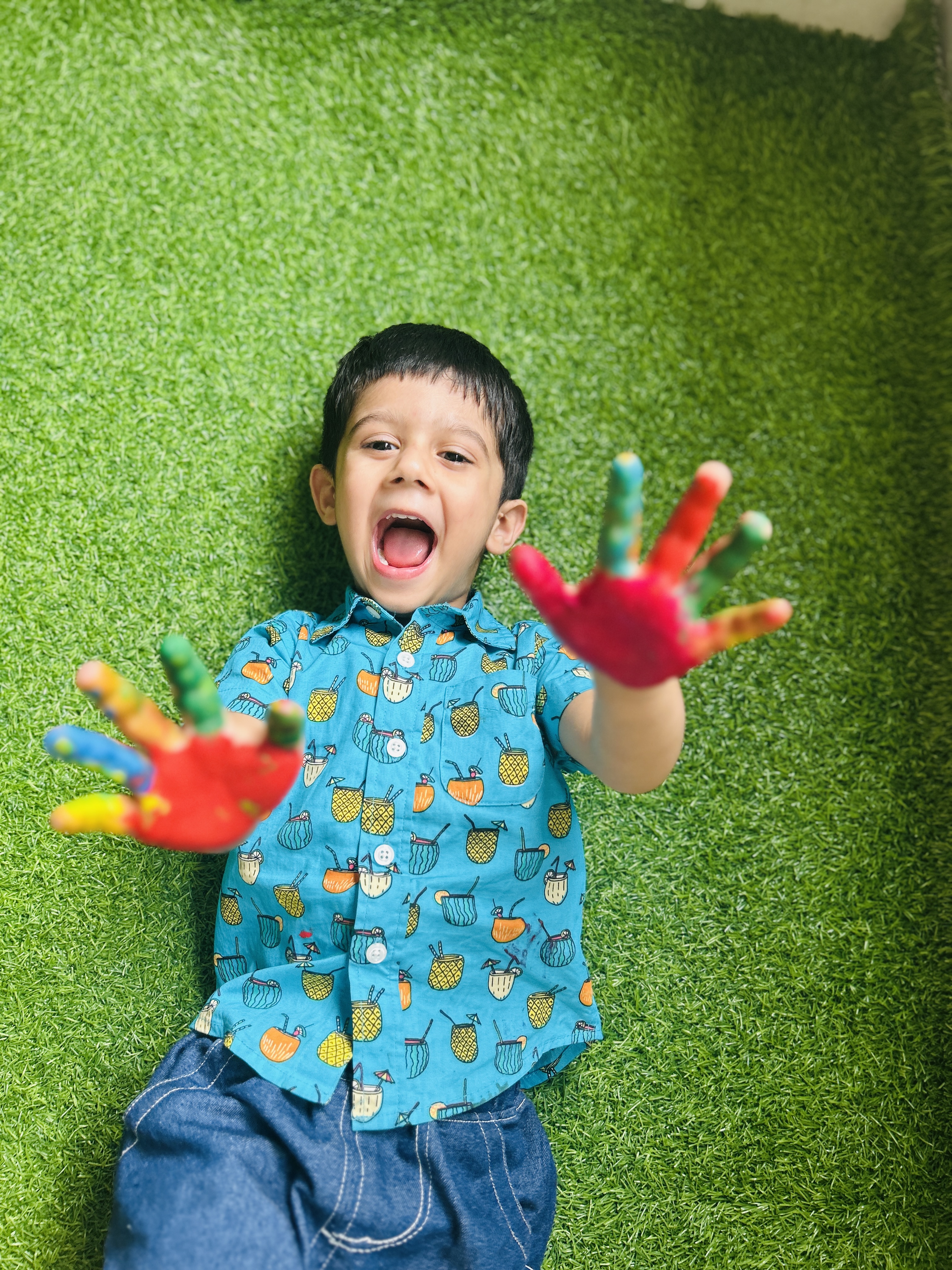 A happy child lying on grass with bright, painted hands raised.
